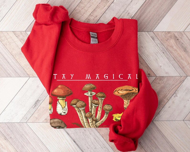 Op de afbeelding: Rood sweatshirt met een witte afbeelding van paddenstoelen en de tekst "STAY MAGICAL".