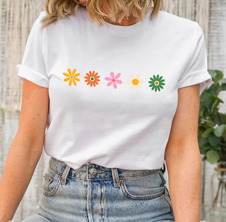 May include: White t-shirt with a row of five colorful flowers printed on the front. The flowers are yellow, orange, pink, white, and green.