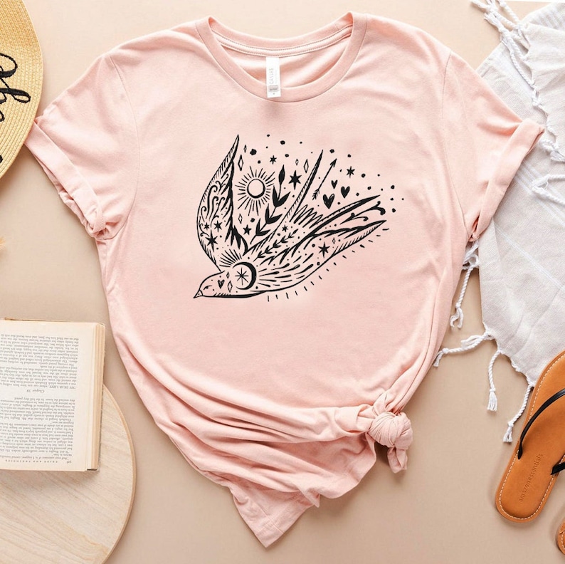 May include: A pink t-shirt featuring a black graphic of a stylised bird with floral and star accents.