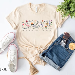 May include: A beige t-shirt with a floral design and the text "Grow Positive Thoughts". The shirt is paired with denim shorts, white sneakers, and a pair of sunglasses.