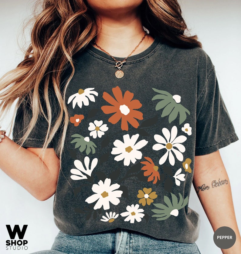 May include: Dark gray t-shirt with a floral design. The graphic features white, orange, green, and yellow flowers. The shirt has the text "W SHOP STUDIO" at the bottom left and "PEPPER" at the bottom right.