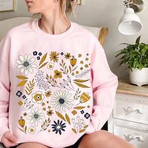 May include: A pink sweatshirt with a floral print featuring yellow, white, and blue flowers. The flowers are arranged in a circular pattern on the front of the sweatshirt.