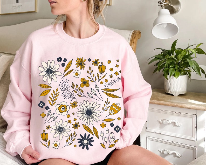 May include: A pink sweatshirt with a floral print featuring yellow, white, and blue flowers. The flowers are arranged in a circular pattern on the front of the sweatshirt.