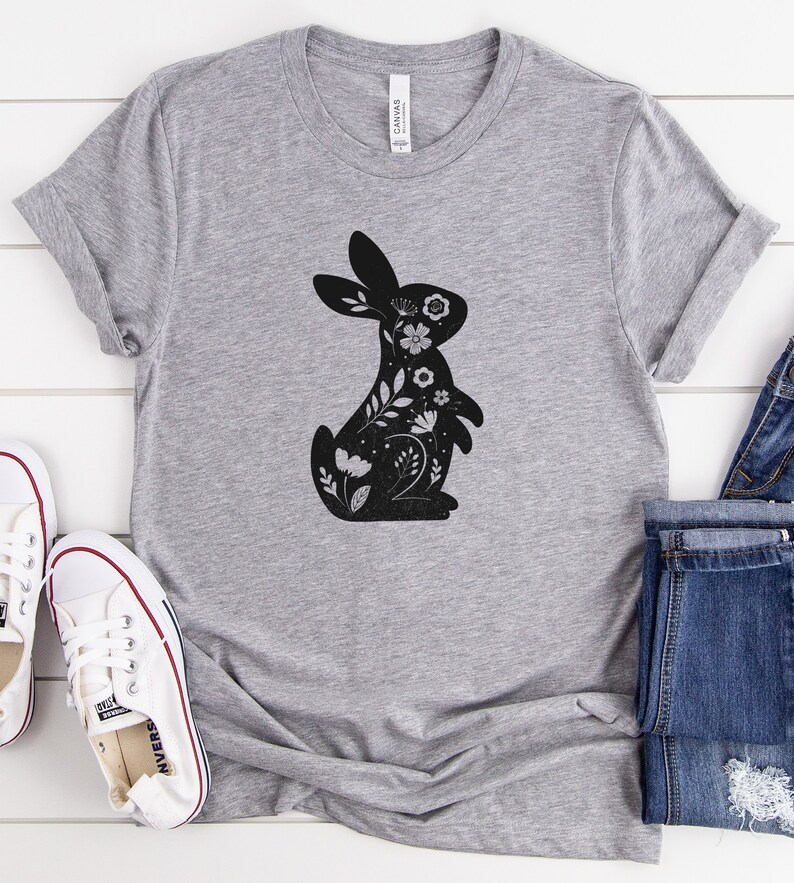 K&ouml;nnte beinhalten: Ein graues T-Shirt mit einer schwarzen Silhouette eines Kaninchens mit floralen Details.
