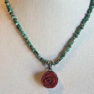 May include: A necklace featuring a strand of small, irregular green beads. A red rose pendant hangs from the necklace. The necklace is displayed on a white mannequin bust.