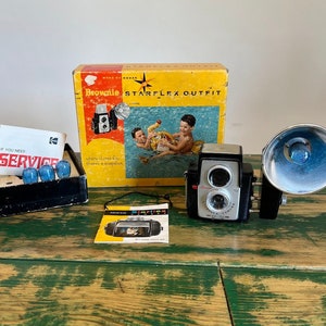May include: A vintage Kodak Brownie Starflex Outfit camera with a flash attachment. The camera is black with a silver lens and a red "Brownie" logo. The box is yellow with red and blue accents and features a picture of two children in a swimming pool. The text on the box reads "Kodak Brownie Starflex Outfit".