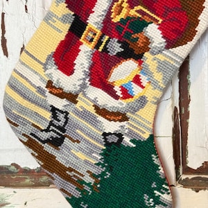May include: Needlepoint Christmas stocking featuring Santa Claus with a red suit, black belt, and a bag of toys. The stocking has a green bottom with a tree design and a yellow background. The design is detailed with various colors.