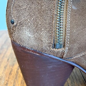 May include: Close-up of a brown suede boot with a dark brown heel. The boot features a metal zipper and decorative stitching. The suede has a textured appearance.