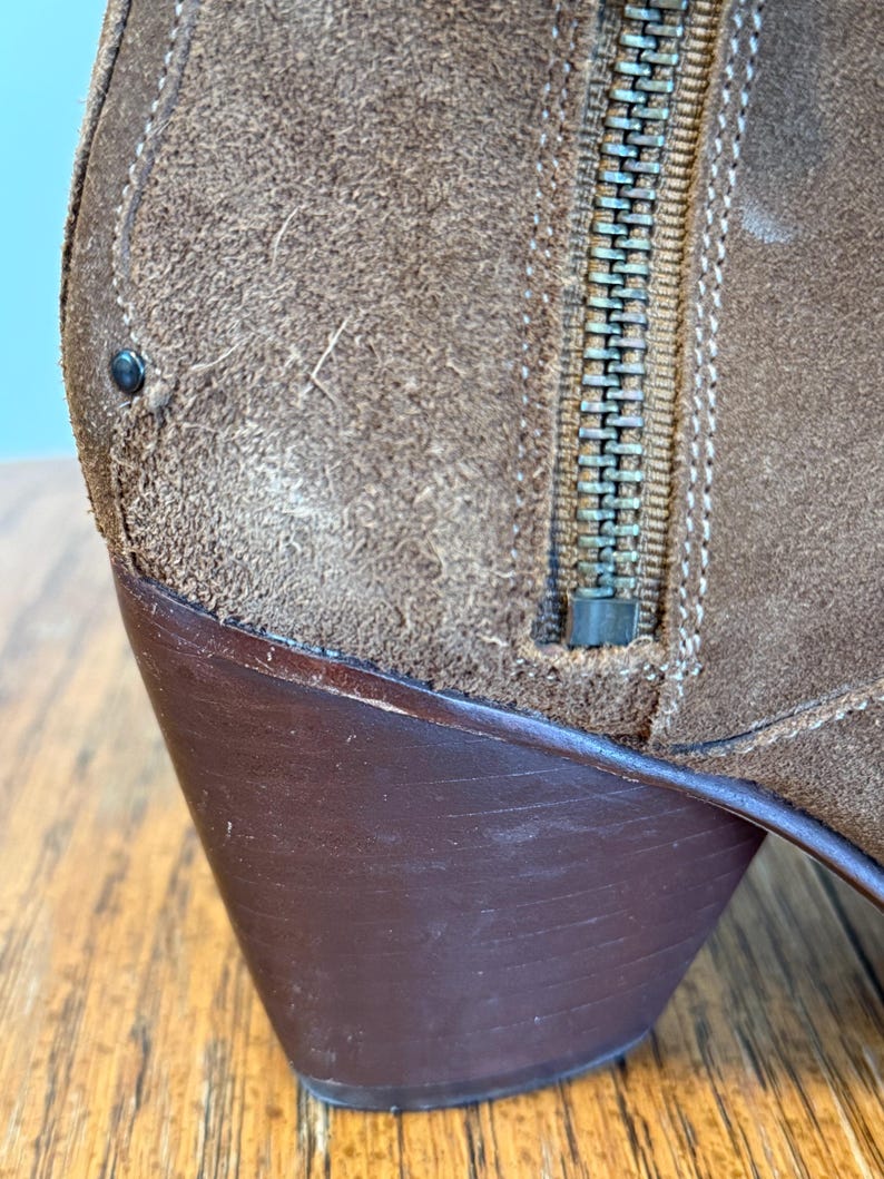 May include: Close-up of a brown suede boot with a dark brown heel. The boot features a metal zipper and decorative stitching. The suede has a textured appearance.