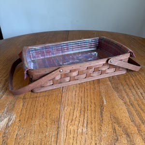 May include: A rectangular brown woven basket with a plaid fabric lining. The basket has two handles and is sitting on a wooden table. The basket is empty.