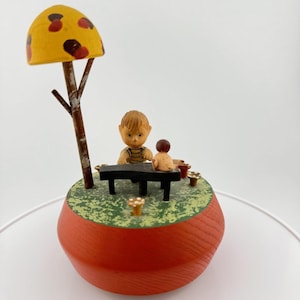 Puede incluir: Una caja de música de madera con un niño y una niña jugando bajo un árbol de hongo amarillo. La base de la caja de música es roja y verde.