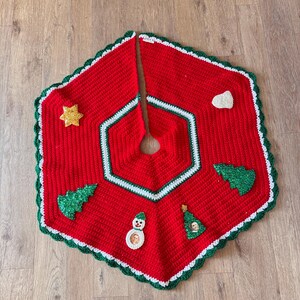 May include: A handmade red crochet Christmas tree skirt with green and white trim. The skirt features appliqued Christmas tree and star designs, along with a snowman. The skirt is designed to go around the base of a Christmas tree.