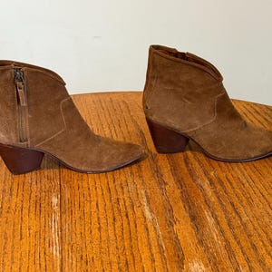 May include: A pair of brown suede ankle boots with pointed toes and stacked heels. Each boot features a side zipper and a V-shaped topline. The boots are displayed on a wooden surface.