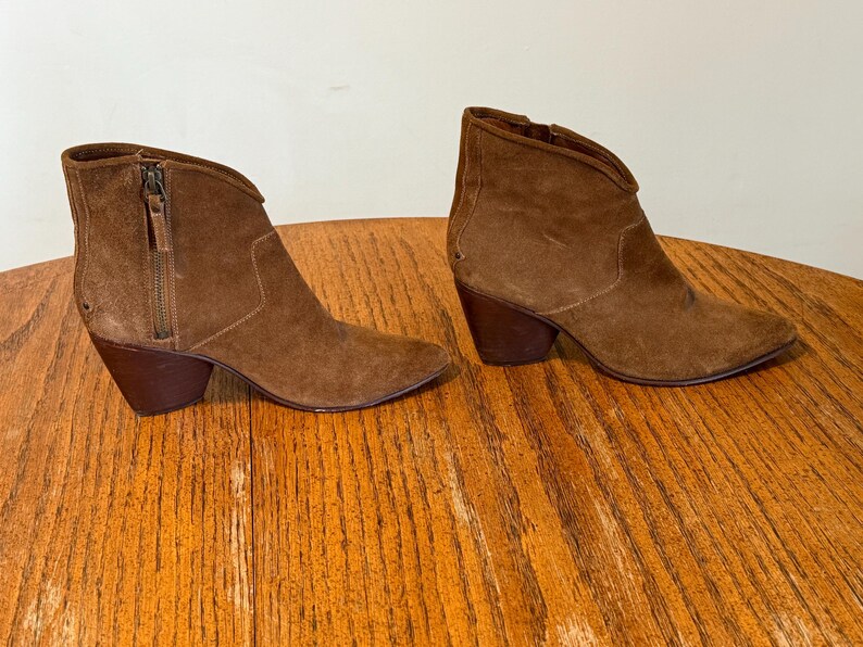 May include: A pair of brown suede ankle boots with pointed toes and stacked heels. Each boot features a side zipper and a V-shaped topline. The boots are displayed on a wooden surface.