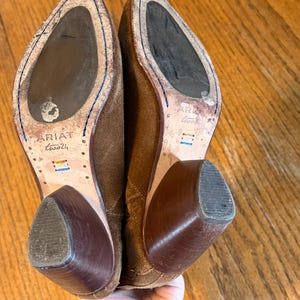 May include: A pair of brown suede Ariat boots with a worn appearance. The soles are visible, showing the Ariat logo and size. The heels are a darker brown, and the boots are held up by a hand.