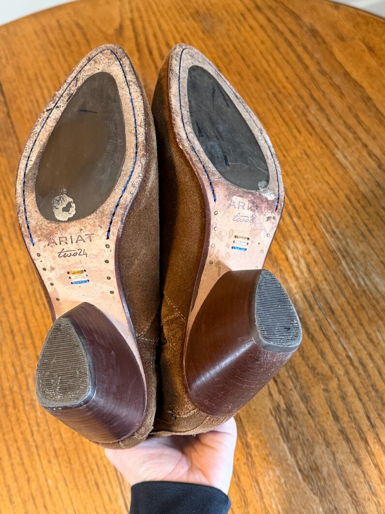 May include: A pair of brown suede Ariat boots with a worn appearance. The soles are visible, showing the Ariat logo and size. The heels are a darker brown, and the boots are held up by a hand.