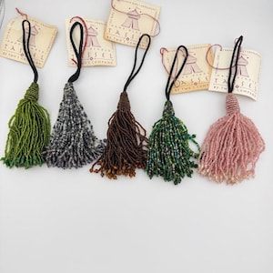 Puede incluir: Cinco borlas decorativas de cuentas en verde, gris, marrón, multicolor y rosa, cada una con un lazo negro para colgar y una etiqueta de papel con la inscripción Tassel by Two's Company. Las borlas presentan intrincados trabajos de cuentas.