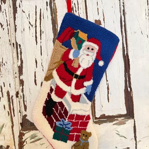 Puede incluir: Un calcetín navideño con un diseño de Papá Noel. El calcetín presenta una parte superior azul, un Papá Noel rojo y blanco, una chimenea y un osito de peluche. El calcetín está hecho de un material texturizado y tiene un lazo rojo para colgar.