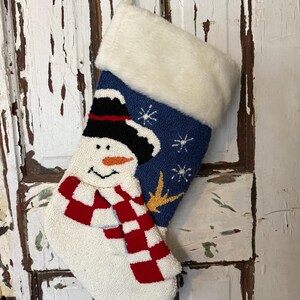 Puede incluir: Un calcetín navideño con un diseño de muñeco de nieve. El calcetín presenta un muñeco de nieve blanco con una bufanda a cuadros rojos y blancos, un sombrero negro y una nariz naranja. La parte superior es azul con copos de nieve blancos y una estrella amarilla.