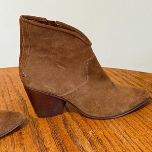 May include: A brown suede ankle boot with a pointed toe and a stacked heel. The boot has a side zipper and a V-shaped topline. The heel measures approximately 3 inches.