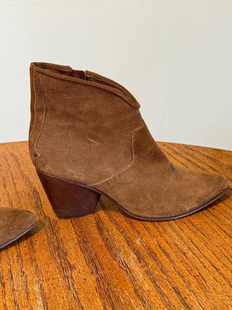 May include: A brown suede ankle boot with a pointed toe and a stacked heel. The boot has a side zipper and a V-shaped topline. The heel measures approximately 3 inches.