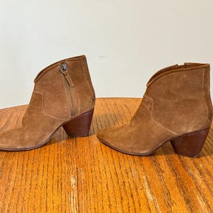 May include: A pair of brown suede ankle boots with a pointed toe and a stacked heel. The boots have a side zipper and a curved topline. The heels are approximately 3 inches high.