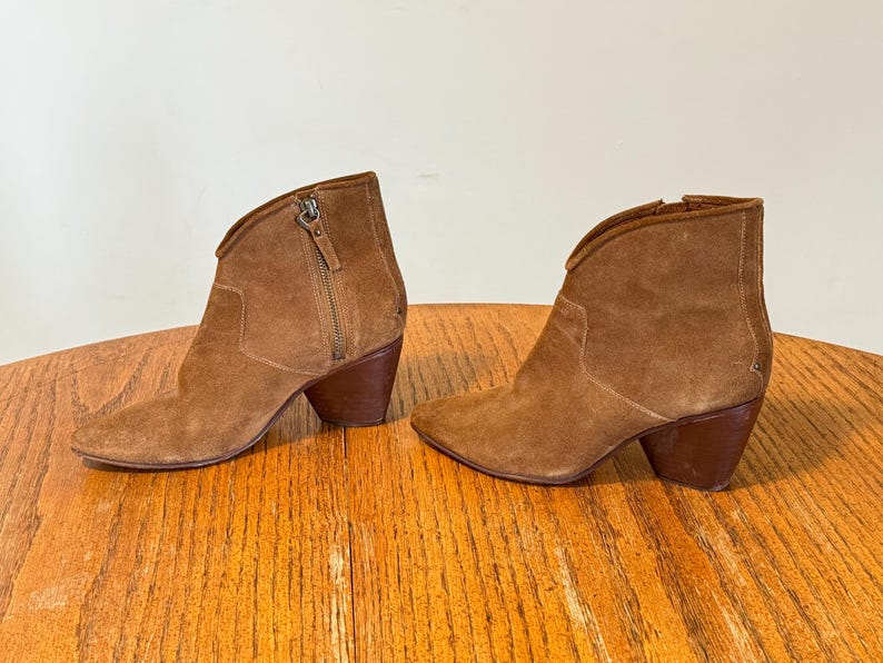 May include: A pair of brown suede ankle boots with a pointed toe and a stacked heel. The boots have a side zipper and a curved topline. The heels are approximately 3 inches high.