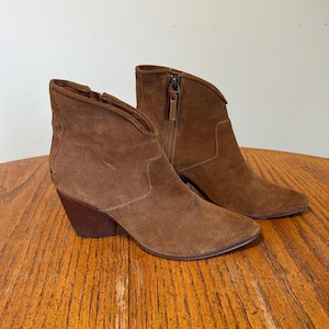 May include: A pair of brown suede ankle boots with a pointed toe and a stacked heel. The boots have a side zipper and a curved topline. The boots are displayed on a wooden surface.