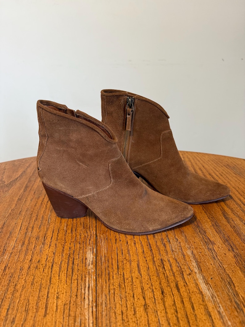 May include: A pair of brown suede ankle boots with a pointed toe and a stacked heel. The boots have a side zipper and a curved topline. The boots are displayed on a wooden surface.