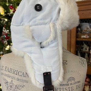 May include: A white winter hat with faux fur trim and ear flaps. The hat features a black button and a black leather strap with a metal buckle. The hat is designed to keep the wearer warm in cold weather.