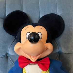 K&ouml;nnte beinhalten: Eine Pl&uuml;sch-Mickey-Mouse-Puppe, die einen blauen Anzug mit einer gelben Weste und einer roten Fliege tr&auml;gt. Die Puppe hat ein breites L&auml;cheln und sitzt auf einem blauen Stuhl.