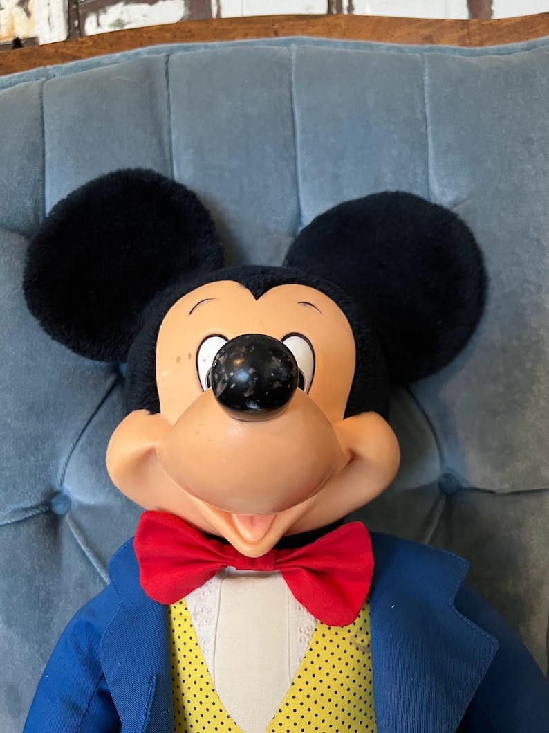K&ouml;nnte beinhalten: Eine Pl&uuml;sch-Mickey-Mouse-Puppe, die einen blauen Anzug mit einer gelben Weste und einer roten Fliege tr&auml;gt. Die Puppe hat ein breites L&auml;cheln und sitzt auf einem blauen Stuhl.