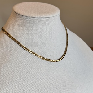 Op de afbeelding: Een goudkleurige ketting met een geweven ontwerp. De ketting wordt getoond op een witte mannequin buste. De ketting heeft een subtiele glans en een klassieke, elegante uitstraling. De achtergrond is neutraal.