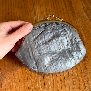 May include: A silver-colored, vintage coin purse with a kiss-lock clasp. The purse has a textured, pleated design and a gold-toned metal frame. The purse is held against a wooden background.