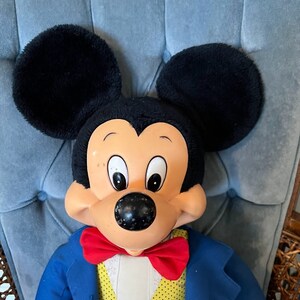 K&ouml;nnte beinhalten: Eine Pl&uuml;sch-Mickey-Mouse-Puppe, die einen blauen Anzug mit einer roten Fliege und eine gelbe Weste mit wei&szlig;en Punkten tr&auml;gt. Die Puppe hat schwarze Ohren und eine schwarze Nase.