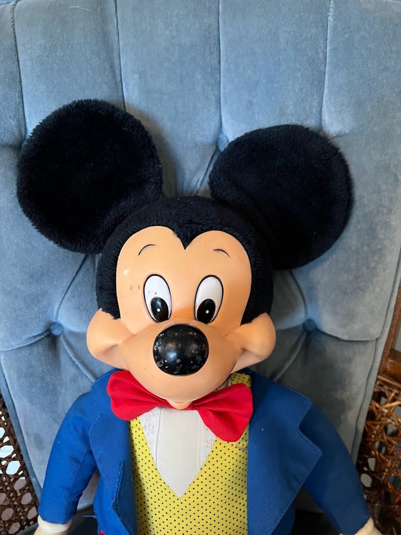 K&ouml;nnte beinhalten: Eine Pl&uuml;sch-Mickey-Mouse-Puppe, die einen blauen Anzug mit einer roten Fliege und eine gelbe Weste mit wei&szlig;en Punkten tr&auml;gt. Die Puppe hat schwarze Ohren und eine schwarze Nase.