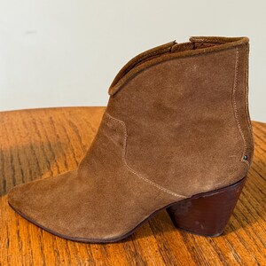May include: A brown suede ankle boot with a pointed toe and a stacked heel. The boot has a V-shaped topline and a side zipper. The heel measures approximately 3 inches.