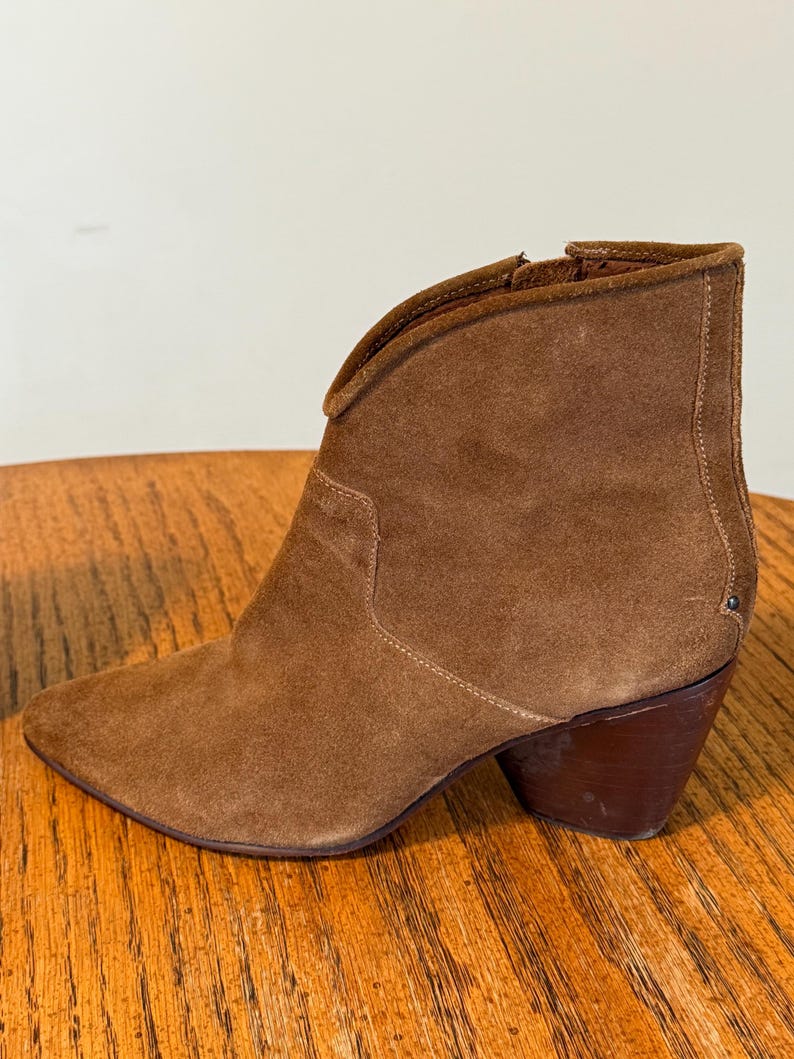 May include: A brown suede ankle boot with a pointed toe and a stacked heel. The boot has a V-shaped topline and a side zipper. The heel measures approximately 3 inches.