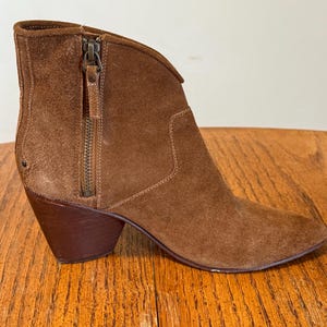 May include: A brown suede ankle boot with a pointed toe and a stacked heel. The boot features a side zipper and a decorative pull tab. The heel measures approximately 3 inches.
