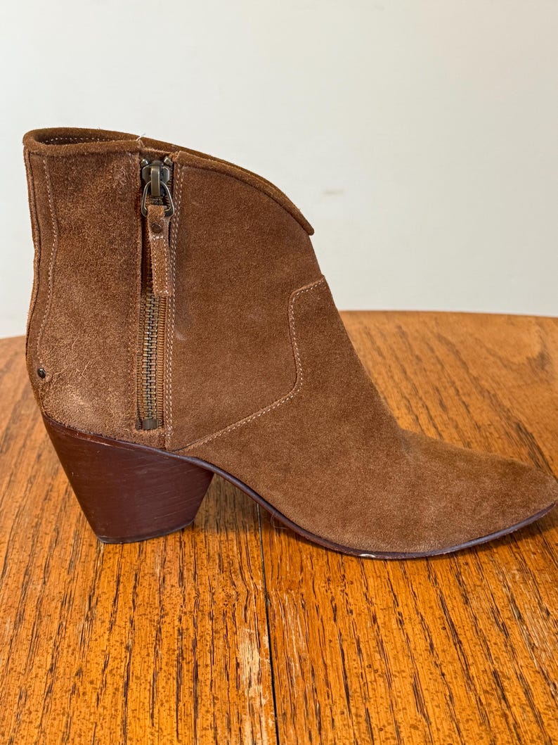 May include: A brown suede ankle boot with a pointed toe and a stacked heel. The boot features a side zipper and a decorative pull tab. The heel measures approximately 3 inches.