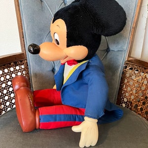 K&ouml;nnte beinhalten: Eine gro&szlig;e Pl&uuml;sch-Mickey-Mouse-Puppe, die einen blauen Anzug mit roten Hosen und einer gelben Fliege tr&auml;gt. Die Puppe sitzt auf einem Stuhl mit blauer Stoffpolsterung.