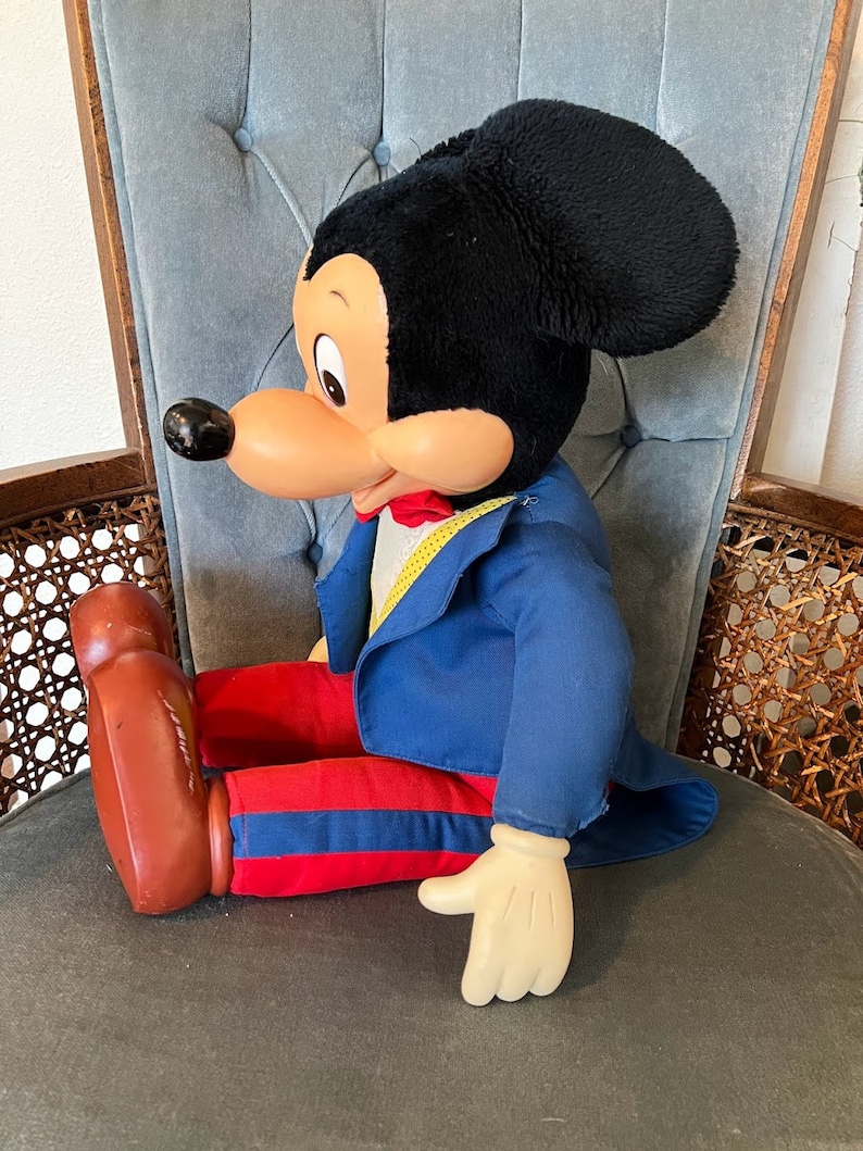 K&ouml;nnte beinhalten: Eine gro&szlig;e Pl&uuml;sch-Mickey-Mouse-Puppe, die einen blauen Anzug mit roten Hosen und einer gelben Fliege tr&auml;gt. Die Puppe sitzt auf einem Stuhl mit blauer Stoffpolsterung.