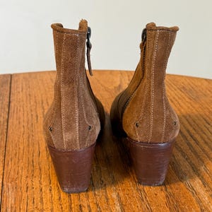 May include: A pair of brown suede ankle boots with a stacked heel. The boots have a zipper on the side and decorative stitching. The heels are approximately 3 inches tall.