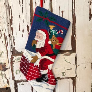 Puede incluir: Un calcetín navideño con un diseño colorido de Papá Noel descendiendo por una chimenea de ladrillos rojos. El calcetín tiene un fondo azul y una cinta roja para colgar. También aparece un osito de peluche. Hecho de un material texturizado.