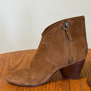 May include: Brown suede ankle boot with a side zipper and a stacked heel. The boot has a pointed toe and a low, angled heel. The suede has a soft, textured appearance. The zipper is silver-toned.