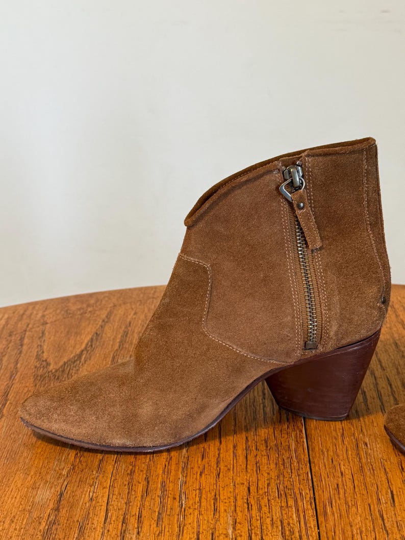 May include: Brown suede ankle boot with a side zipper and a stacked heel. The boot has a pointed toe and a low, angled heel. The suede has a soft, textured appearance. The zipper is silver-toned.