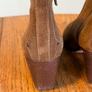 May include: Close-up of a pair of brown suede ankle boots. The boots have a stacked heel and a zipper on the side. The suede is a warm, earthy tone, and the stitching details add texture.