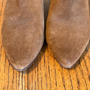 May include: Close-up of two brown suede boots with pointed toes. The boots have a worn, textured appearance, suggesting they are well-loved. The image focuses on the front of the boots, highlighting their shape and material.