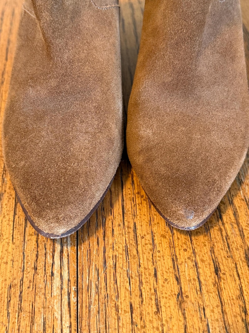 May include: Close-up of two brown suede boots with pointed toes. The boots have a worn, textured appearance, suggesting they are well-loved. The image focuses on the front of the boots, highlighting their shape and material.