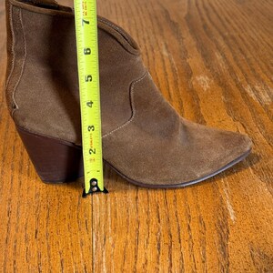 May include: A brown suede ankle boot with a pointed toe and a stacked heel. The boot is approximately 8 inches tall, measured with a yellow tape measure. The boot has a V-shaped topline and a textured panel on the back.
